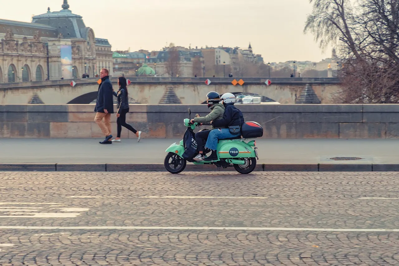 Deux personnes casquées sur un scooter vert, le long d’un quai pavé en ville